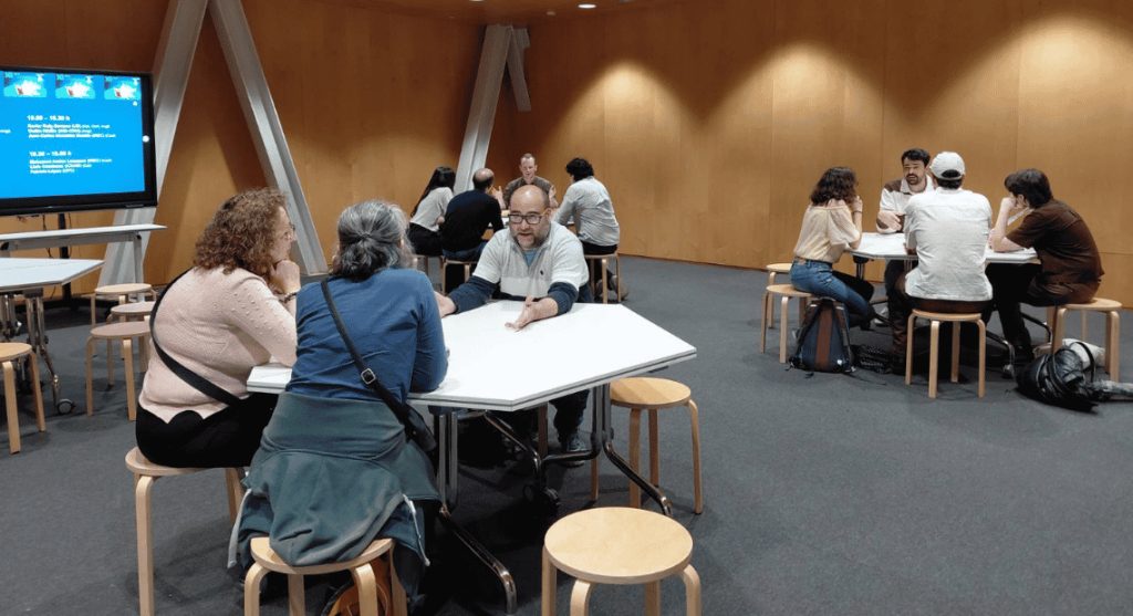 A group of people engaged in small group discussions at white tables during the Festival 10alamenos9 event.