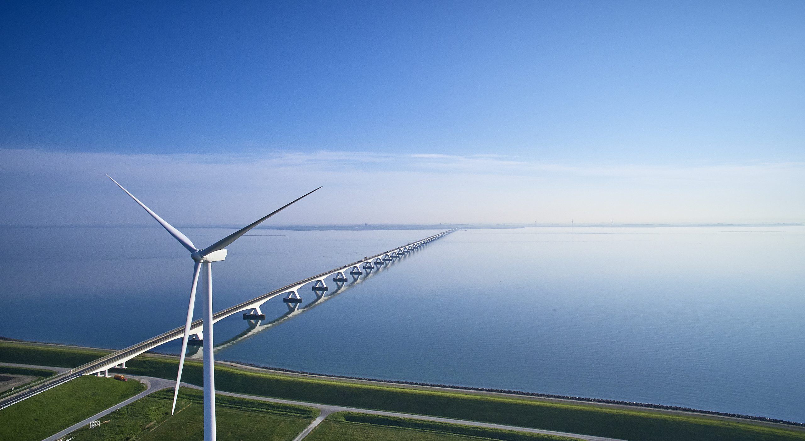 A large wind turbine standing by a coastal bridge, exemplifying IREC’s strategic infrastructures and its mission to provide sustainable energy solutions.