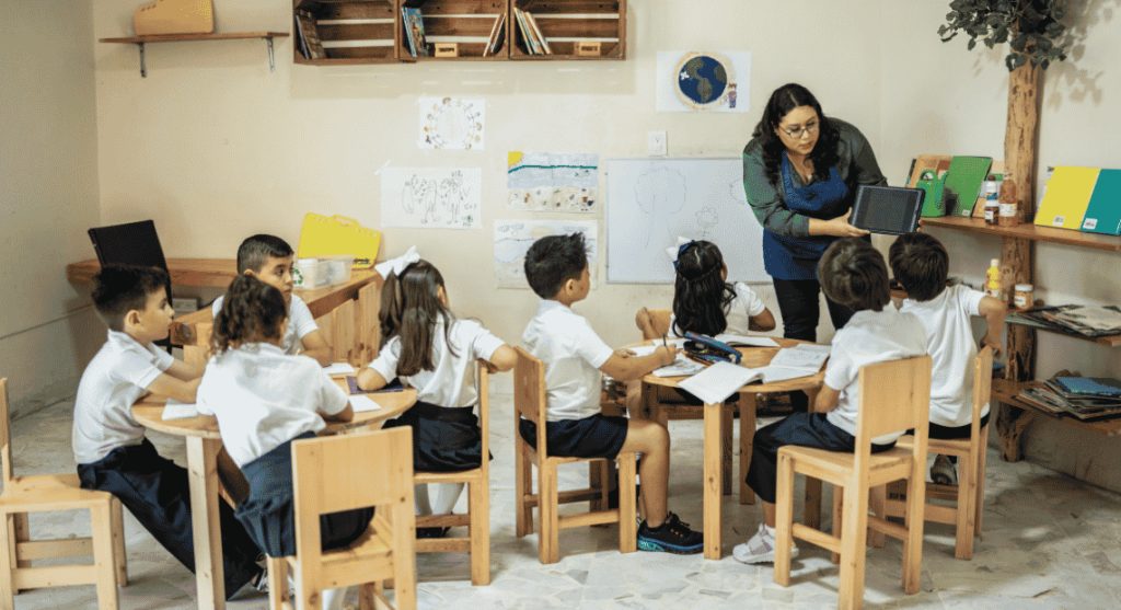 A teacher showing a tablet to a group of young students sitting at wooden tables in a classroom.